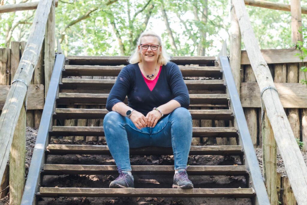 Cat Boersma sitting outdoors on wooden steps in the forest, smiling with openness and warmth, symbolizing guidance and support for expat women through nature therapy.
