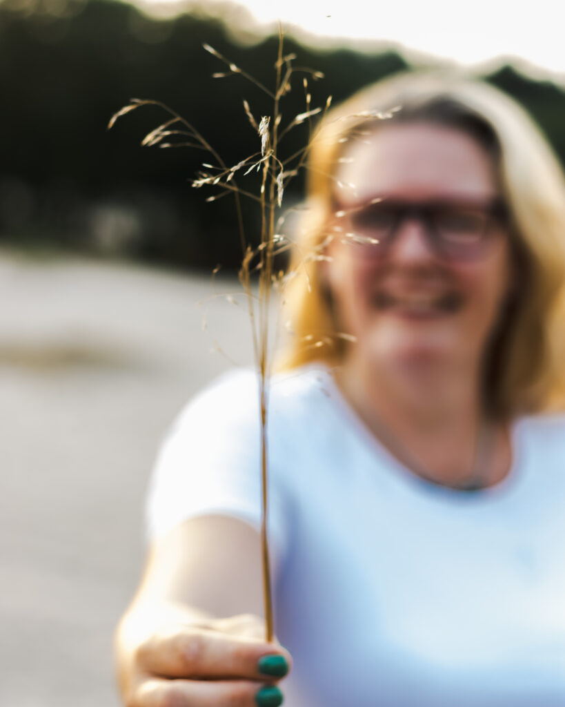 Cat Boersma smiling while holding out a fragile stem of grass, symbolizing connection, resilience, and presence in nature.beauty of nature’s small details.