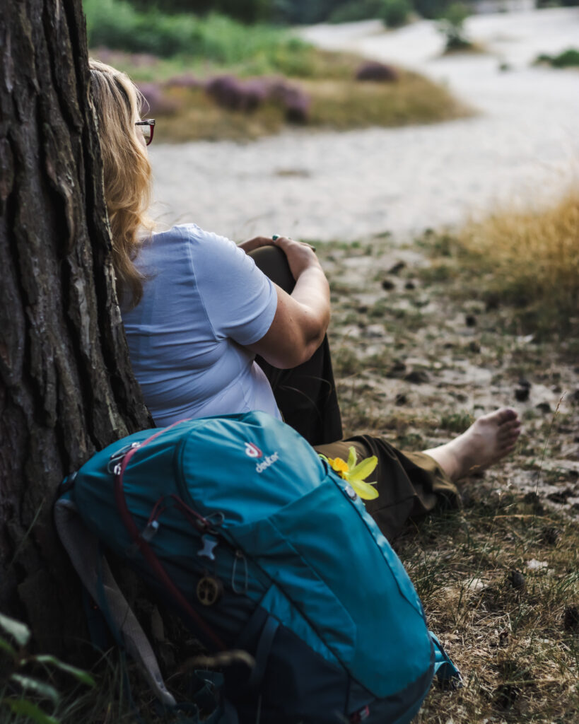 Cat Boersma resting barefoot against a tree with her backpack, finding stillness and support in nature.